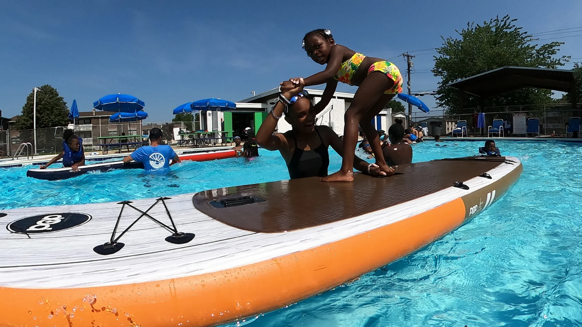 A girl stands with assistance on an inflatable paddleboard in a pool