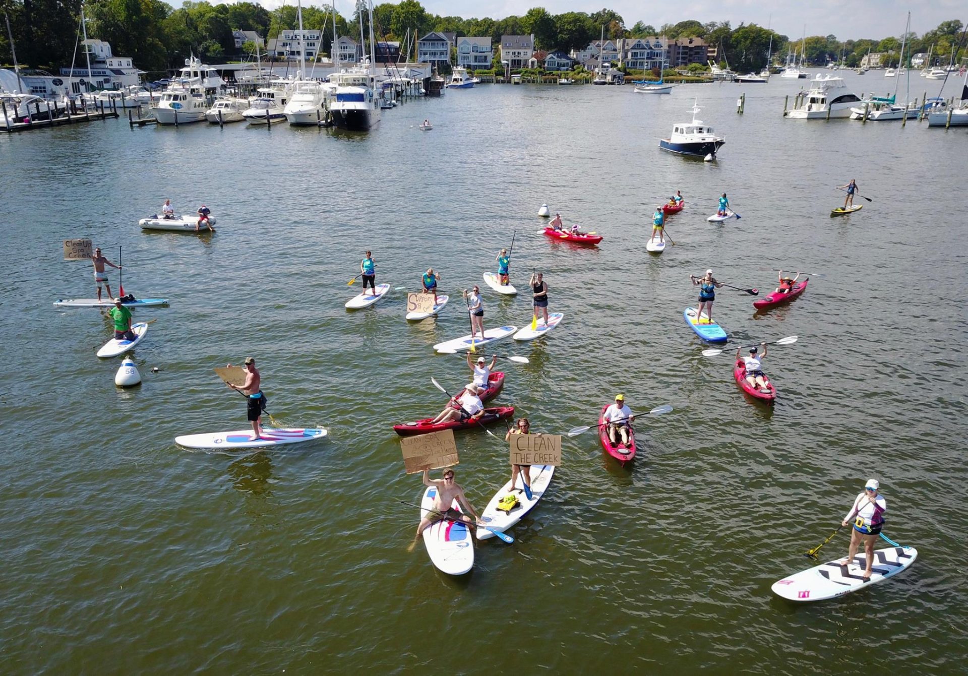 Overhead shot of several stand-up paddlers on the water with signs. One reads "Clean the Creek"