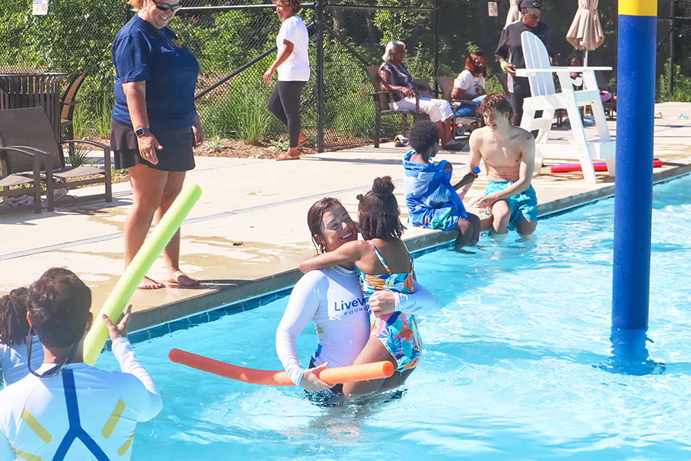 Woman holds child in a pool with a pool noodle in her other hand
