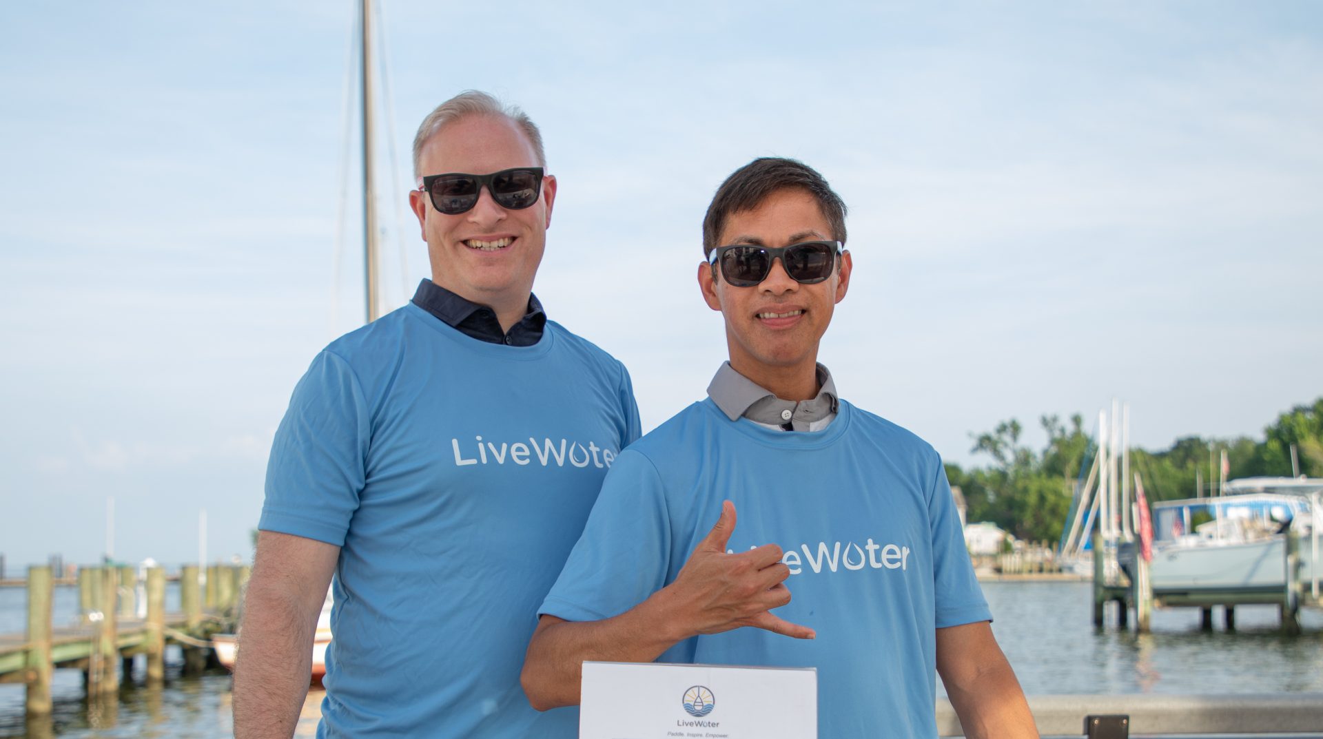 Two men stand smiling, wearing blue LiveWater t-shirts, in front of a waterfront. One is throwing a shaka.