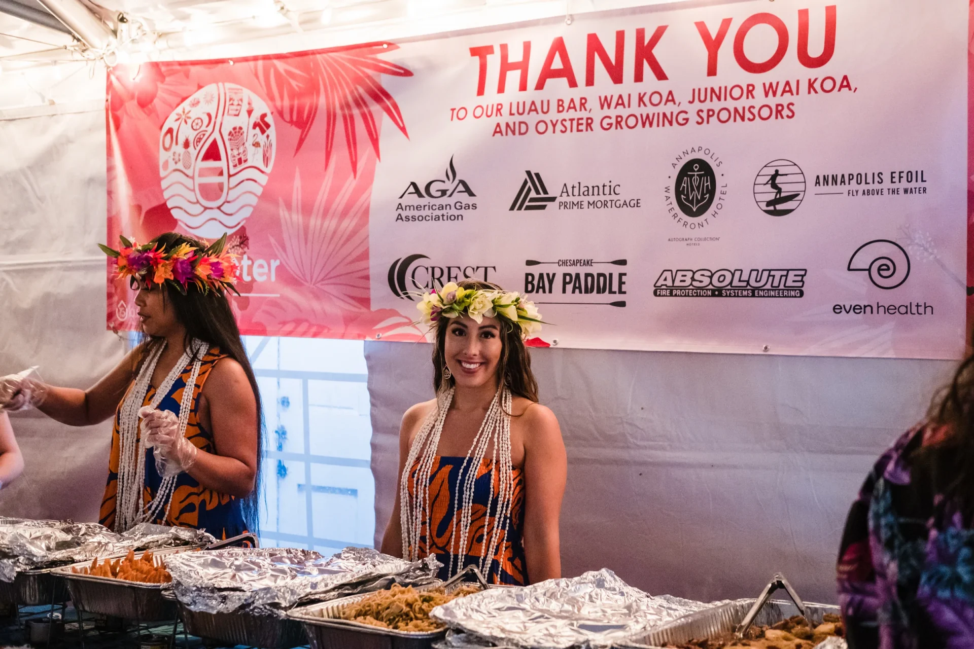 Two women in aloha gear and flower leis behind a serving table full of food