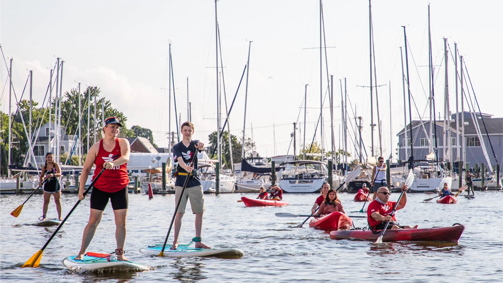 stand-up paddlers and kayakers paddle up a creek with boats in the background