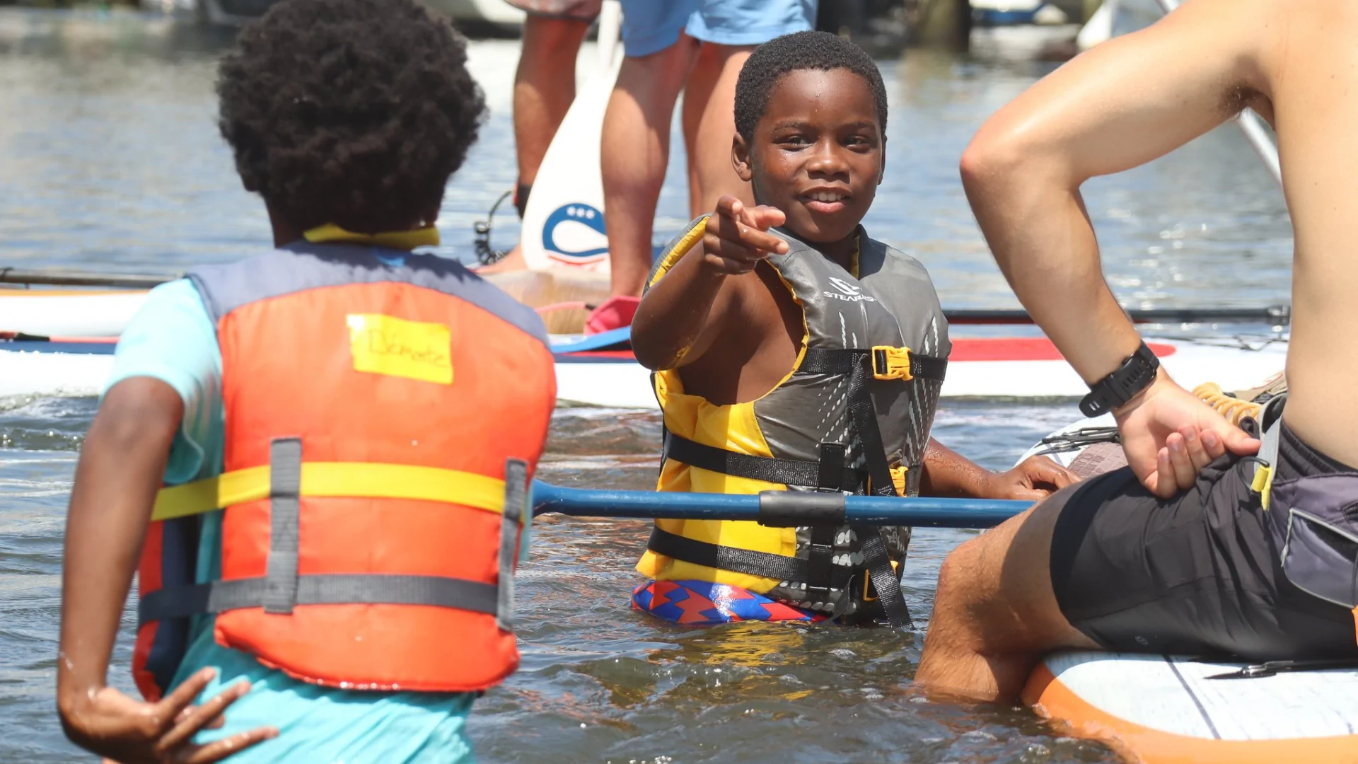 a smiling child stands in the water in a life vest and points toward the camera.