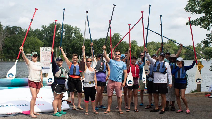 A group holds up their paddles in front of some stand-up boards and the water.