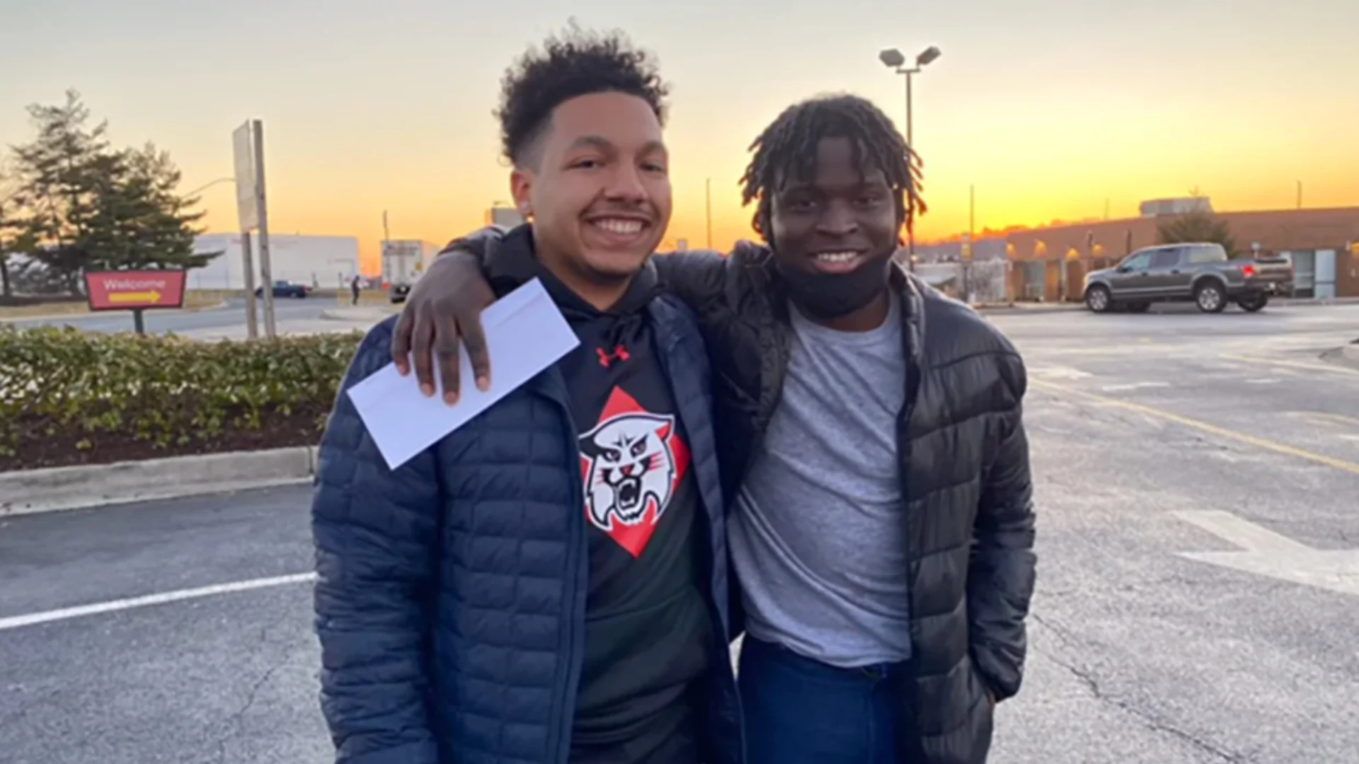 Two men stand in a parking lot with arms around each other and the sun setting behind