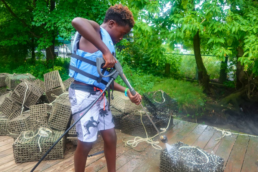 A boy in a life vest sprays dirty oyster cages with a pressure washer.