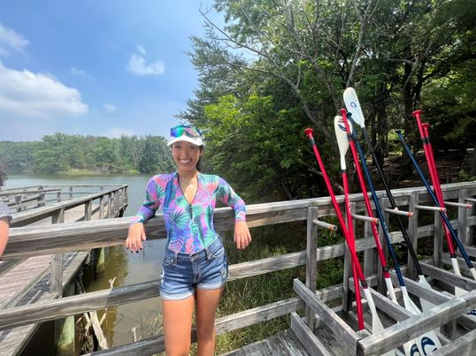 Gaby stands against the railing of a dock at Quiet Waters Park. She is wearing a blue and pink top and denim shorts, with a white cap and sunglasses. Next to her are a row of paddles for kayaks and stand up paddle boards.