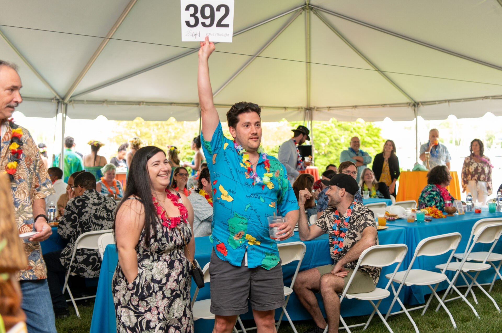 A man dressed in a blue hawaiian print shirt and dark grey shorts holds an auction card up with the number 392. There is a woman in a black and white floral dress standing to his right, with a number of people seated in the background behind him. 