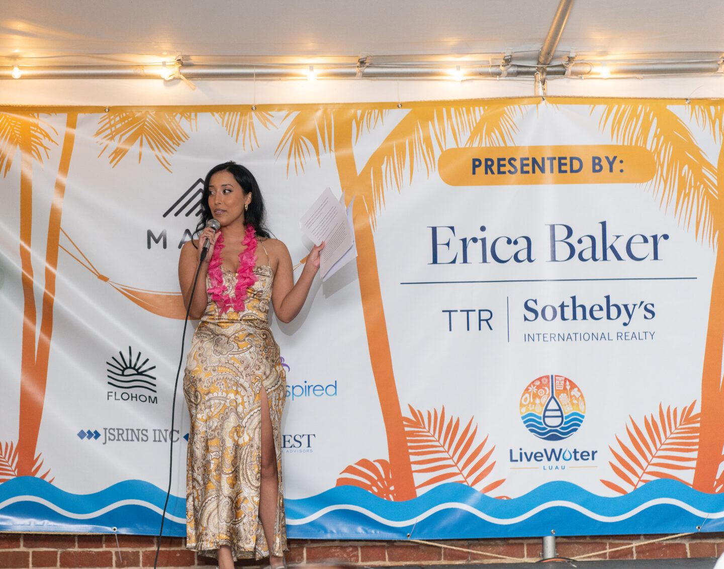 Gaby stands on a stage in front of a backdrop with a microphone in her hand. She is wearing a gold colored dress with swirling patterns on it.