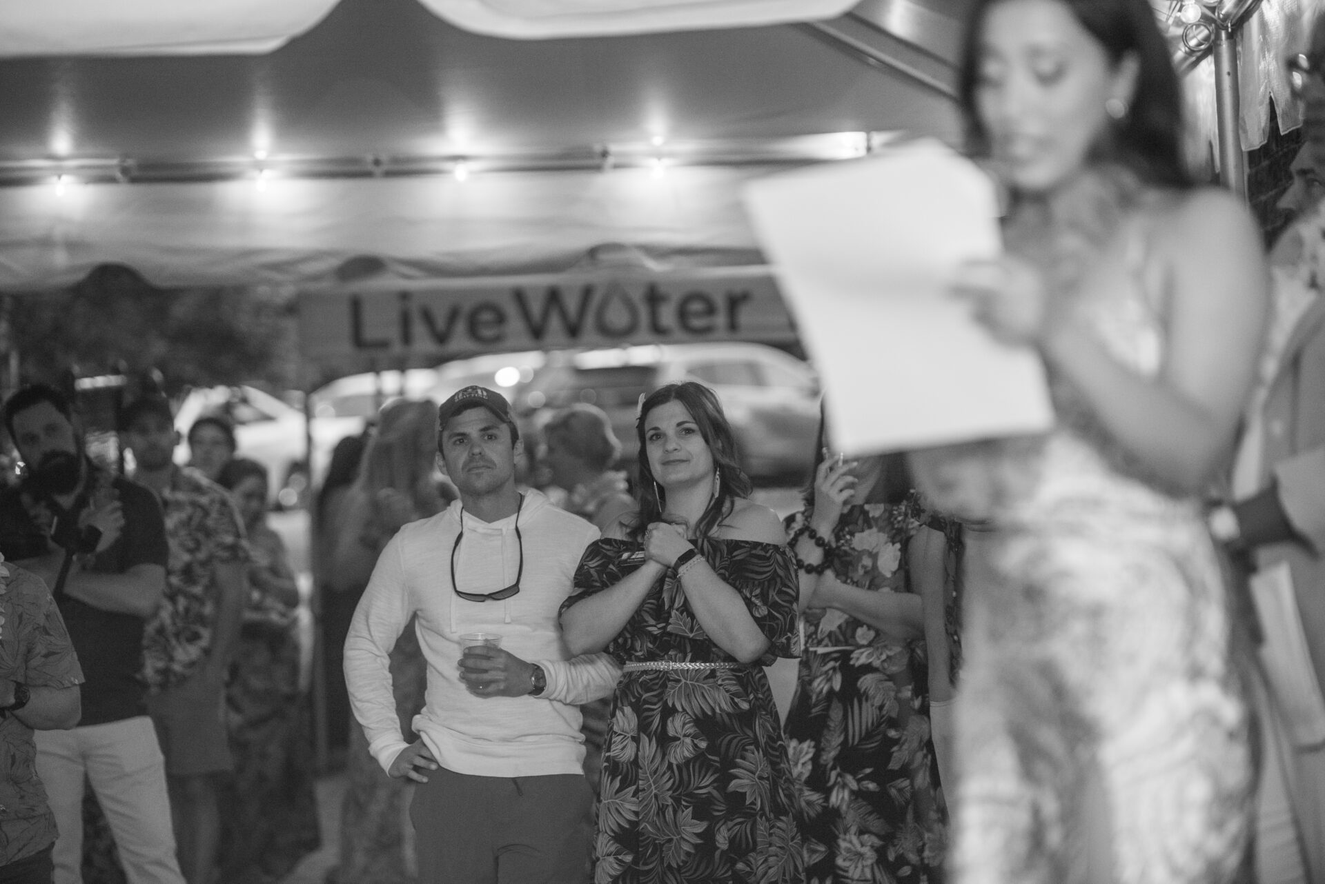 Gaby stands on stage in front of a crowd at the Live Water Luau. She is speaking into a microphone and looking down at her speech notes; three onlookers in the foreground are visibly moved. The photo is in black and white.
