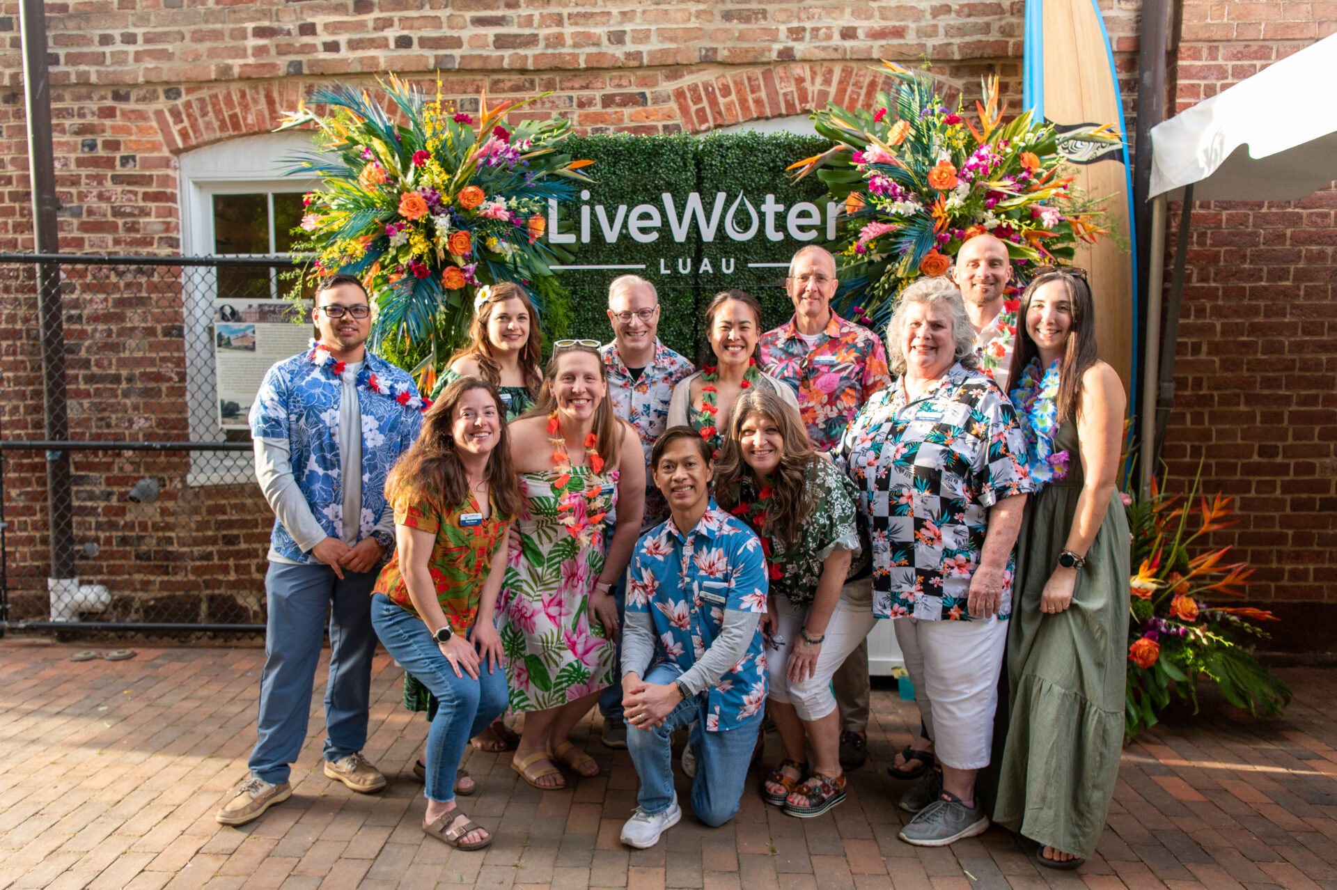 The Live Water Foundation board of directors, all dressed in Hawaiian-themed clothing, pose in front of a green backdrop that says "live water foundation". The display is flanked by floral arrangements.