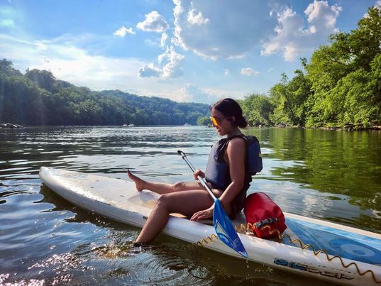 Gaby sits on top of a paddle board in the water. She is surrounded by trees and has one leg dangling in the water.