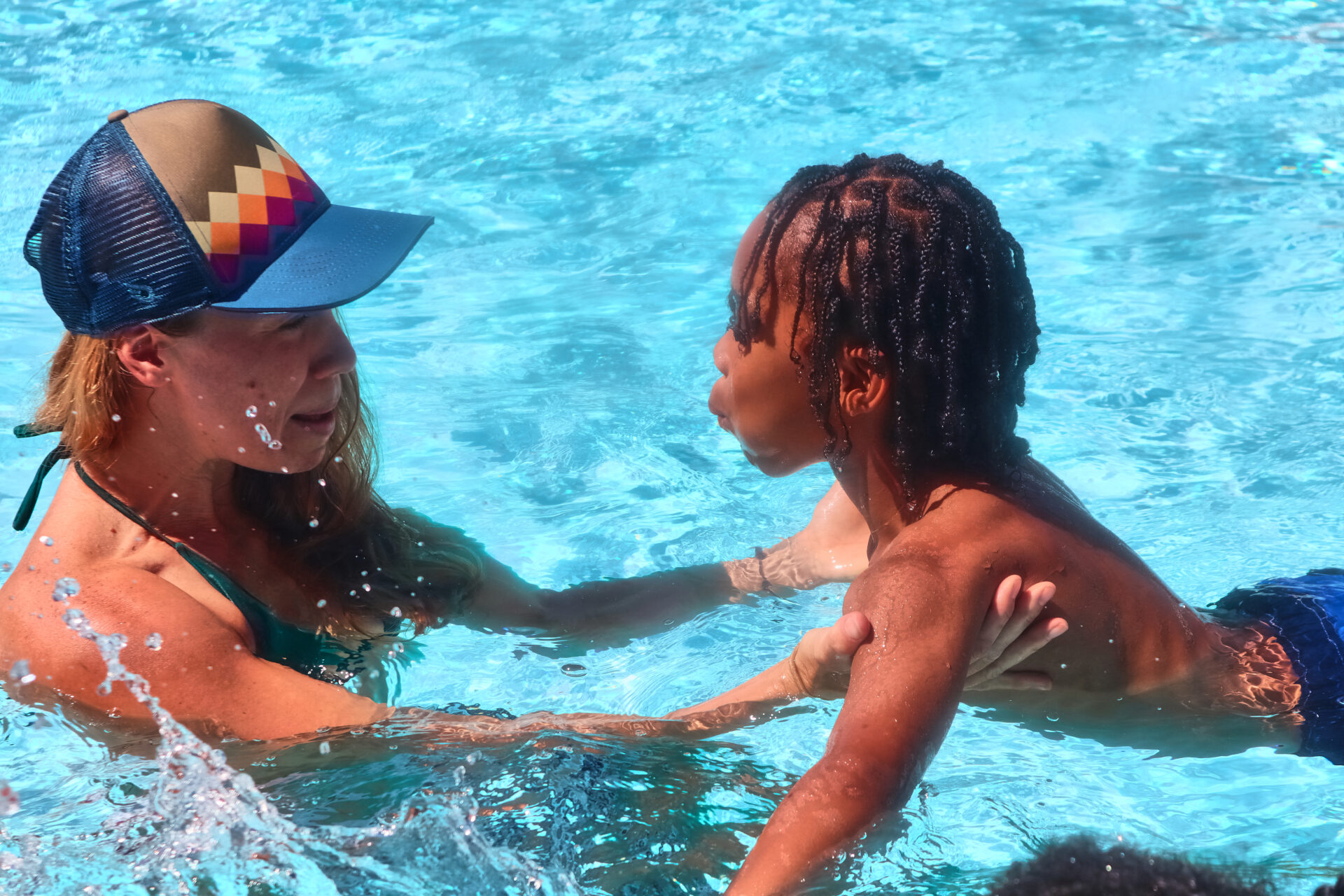 A woman with a blue baseball cap is helping a child swim in a pool. She appears to be showing him breathing techniques.