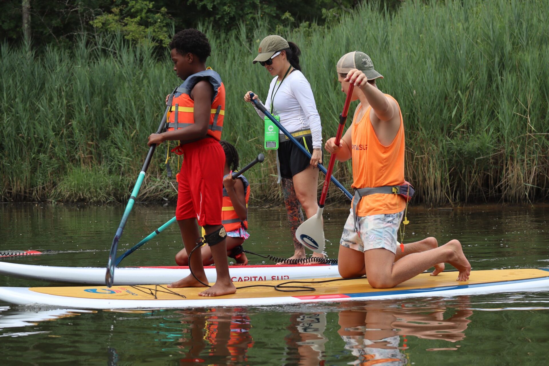 Two adults are teaching two children how to paddle board. There are two paddle boards in the water surrounded by tall grasses. Each paddle board has one adult and one child on it.