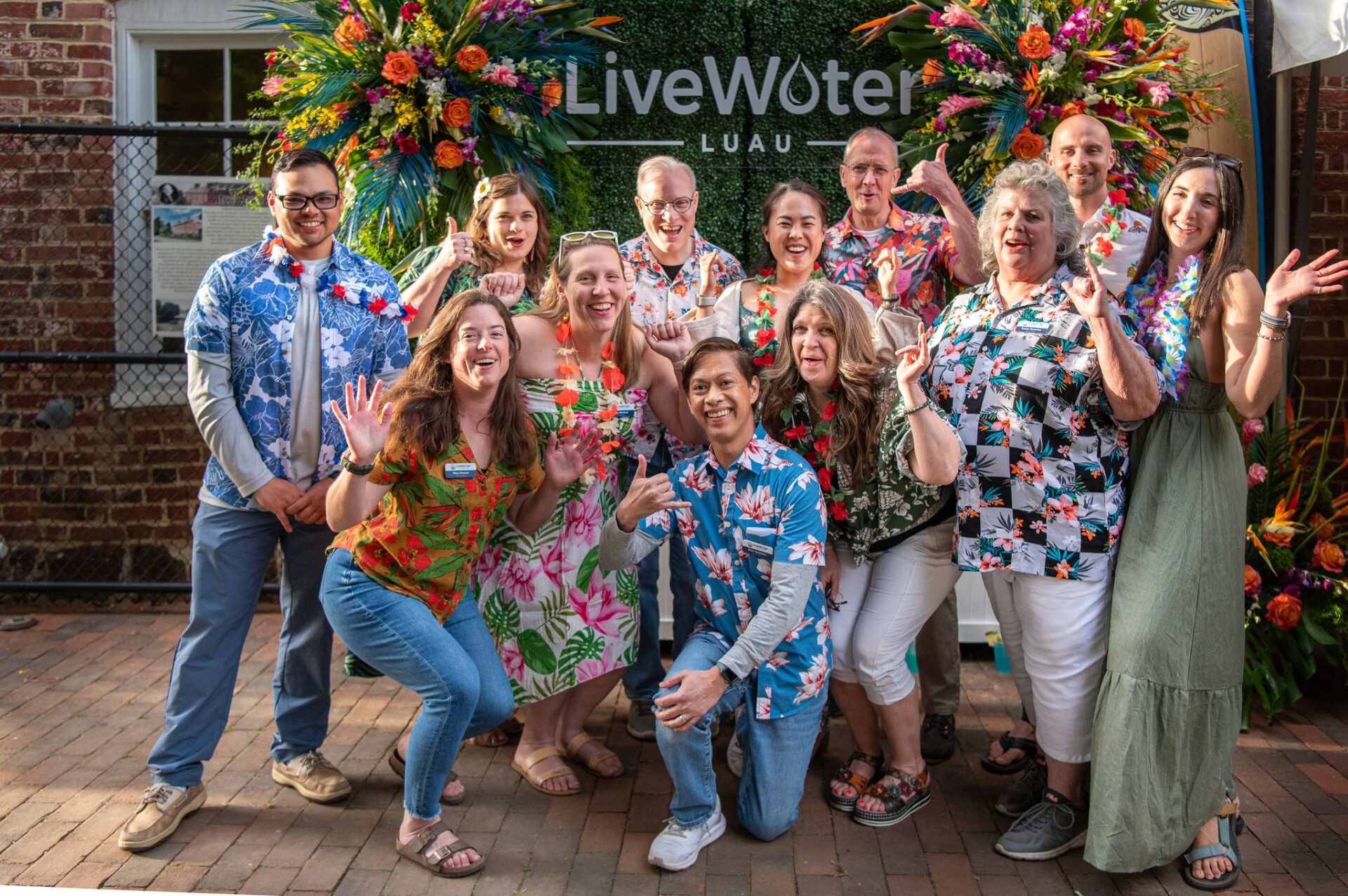 Live Water Foundation board members pose as a group in front of a banner that reads "LiveWater". They are flanked by tropical flowers and wearing luau-themed clothing.